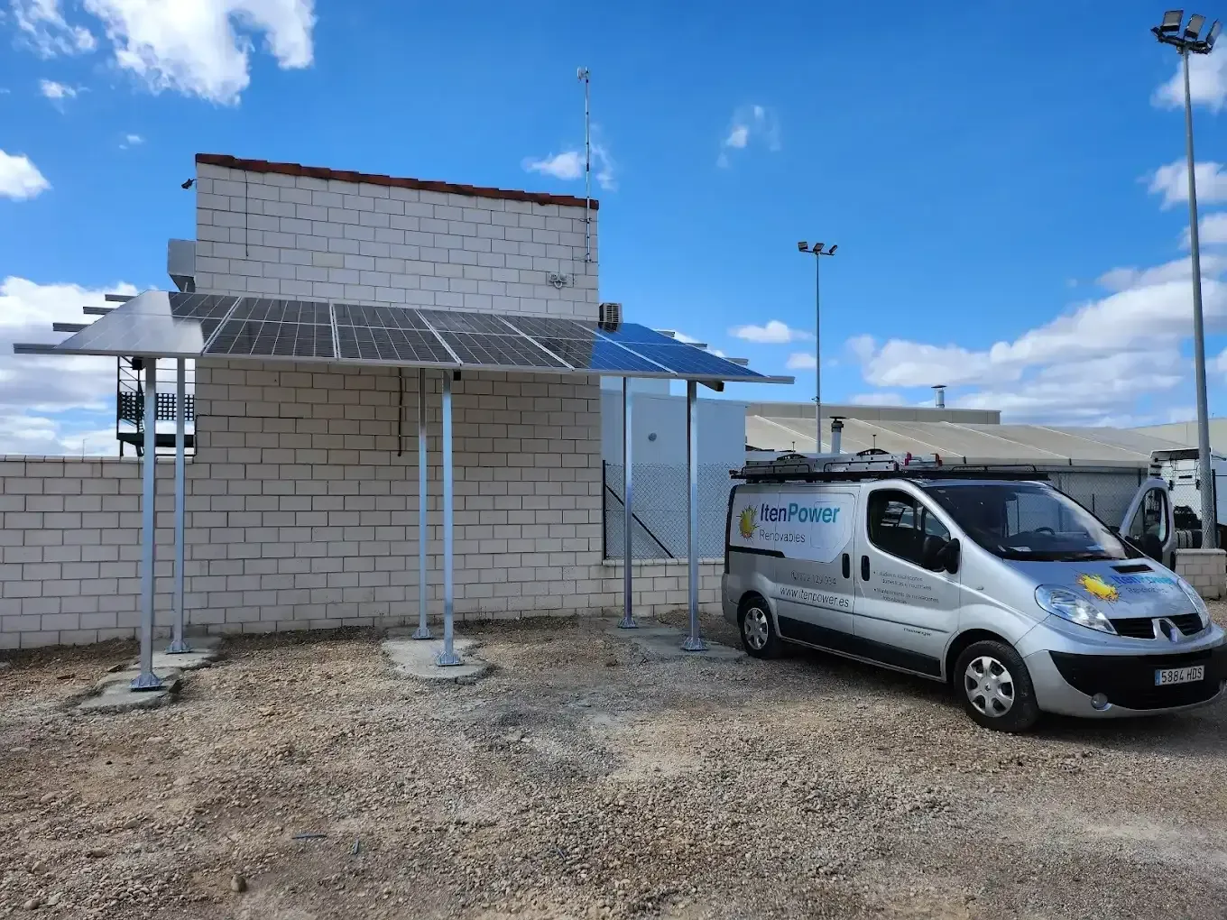Furgoneta blanca aparcada junto a un pequeño edificio de ladrillos bajo un cielo azul en un terreno de grava.