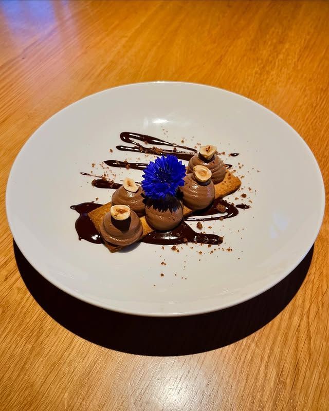 Assiette à dessert nappée de chocolat, composée de petites pâtisseries et décorée d'une fleur bleue, sur une table en bois.