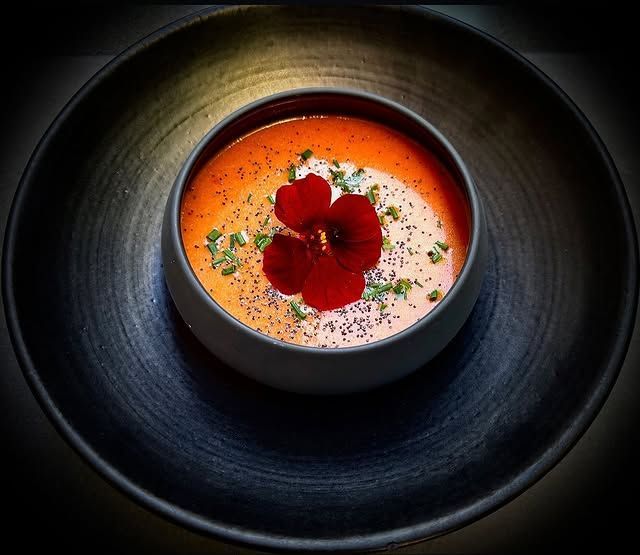 Soupe crémeuse à l'orange servie dans un bol noir, garnie d'une fleur rouge et d'herbes aromatiques sur une assiette foncée.