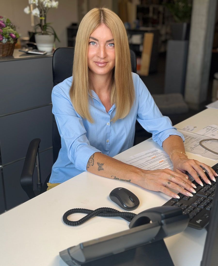 Eine Frau an einem Schreibtisch, die Tastatur und Maus benutzt, ein hellblaues Hemd trägt und in die Kamera lächelt.