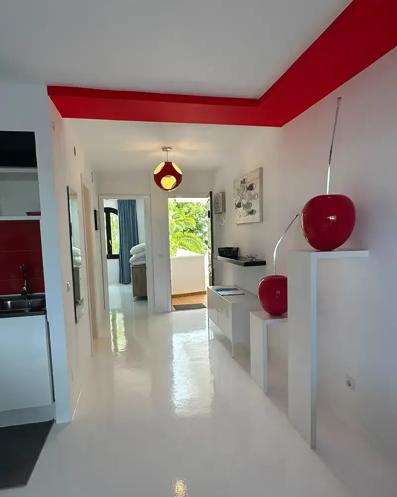Pasillo blanco luminoso con detalles en rojo, lámparas colgantes y vistas a una habitación soleada y al jardín.
