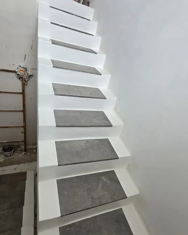 Escalera estrecha blanca con escalones de azulejos grises contra una pared lisa, en el interior.