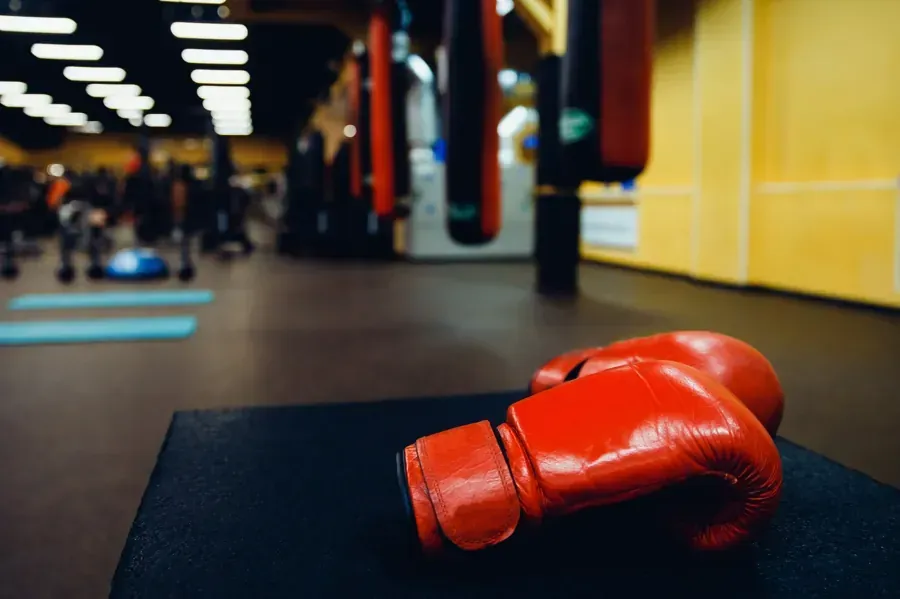 Guantes de boxeo rojos sobre una superficie negra en un gimnasio con sacos de boxeo y figuras borrosas en el fondo.