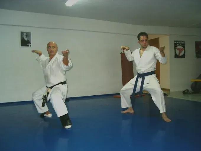 Dos hombres con uniforme de karate practican una postura sobre un tatami azul. Uno lleva cinturón negro y el otro azul.