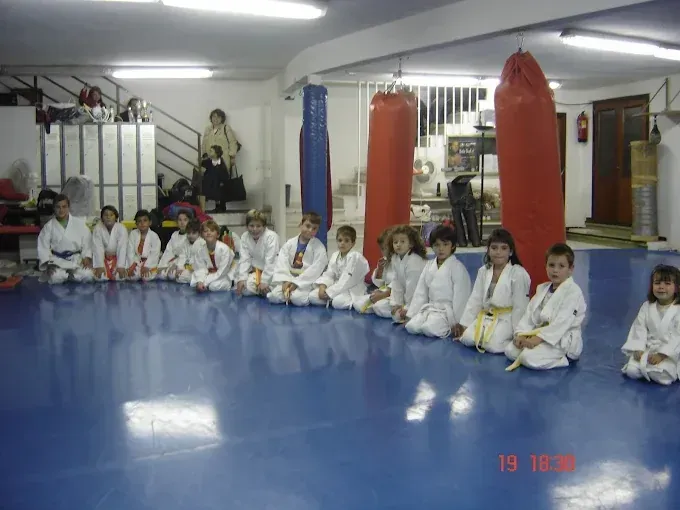 Niños con uniformes de karate blancos se sientan en una colchoneta azul, en una sala de entrenamiento de artes marciales.
