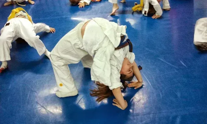 Chica con uniforme de judo blanco haciendo una flexión hacia atrás sobre una colchoneta azul, rodeada de otras personas.
