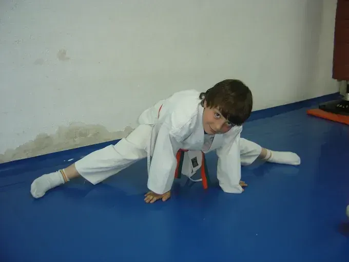 Un joven con uniforme de judo blanco hace el split sobre una colchoneta azul, con las manos en el suelo y sonriendo.