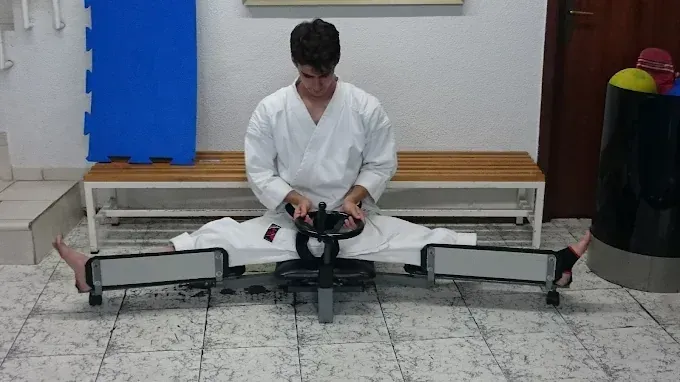 Persona con uniforme de karate usando una máquina de estiramiento, con las piernas bien abiertas. Interior.