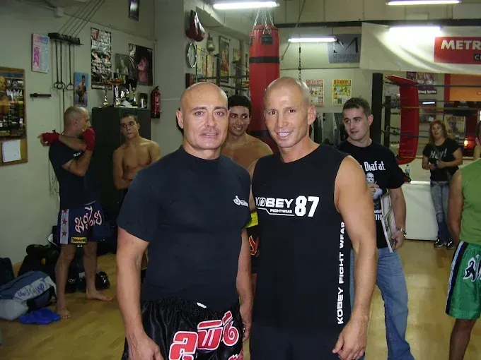 Dos hombres en un gimnasio de Muay Thai, sonriendo, posan para una foto. Otros al fondo.