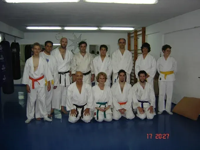 Grupo de personas con uniformes de artes marciales, algunos arrodillados, en un dojo.