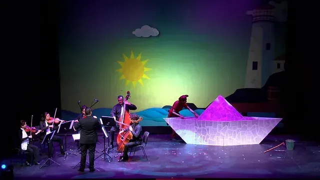 Un grupo de personas está tocando instrumentos en un escenario frente a un barco de papel.