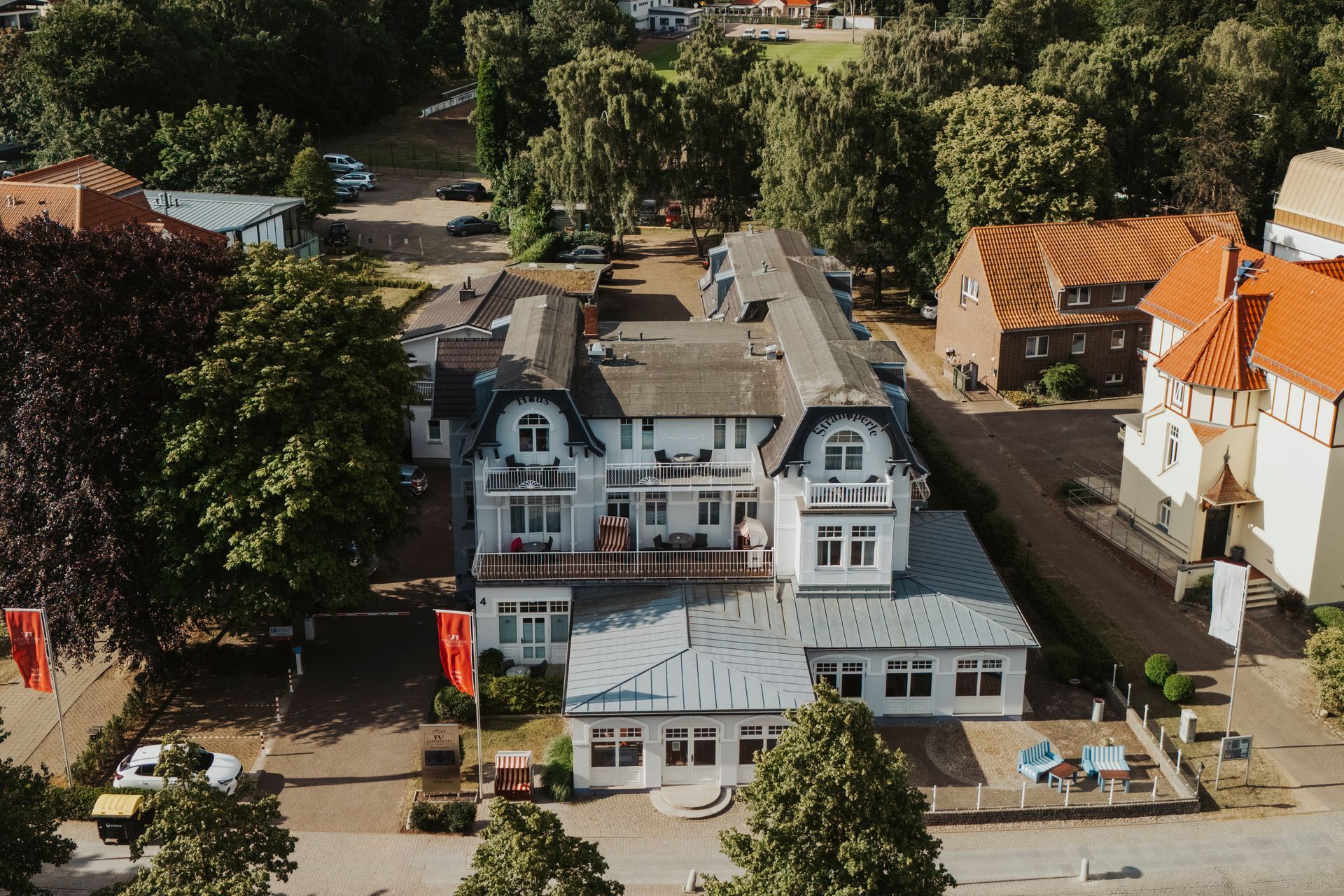 Das hellgraue Gebäude der Strandperle IV Jahreszeiten Kühlungsborn  mit einem Restaurant, Parkplatz und einem Baum.