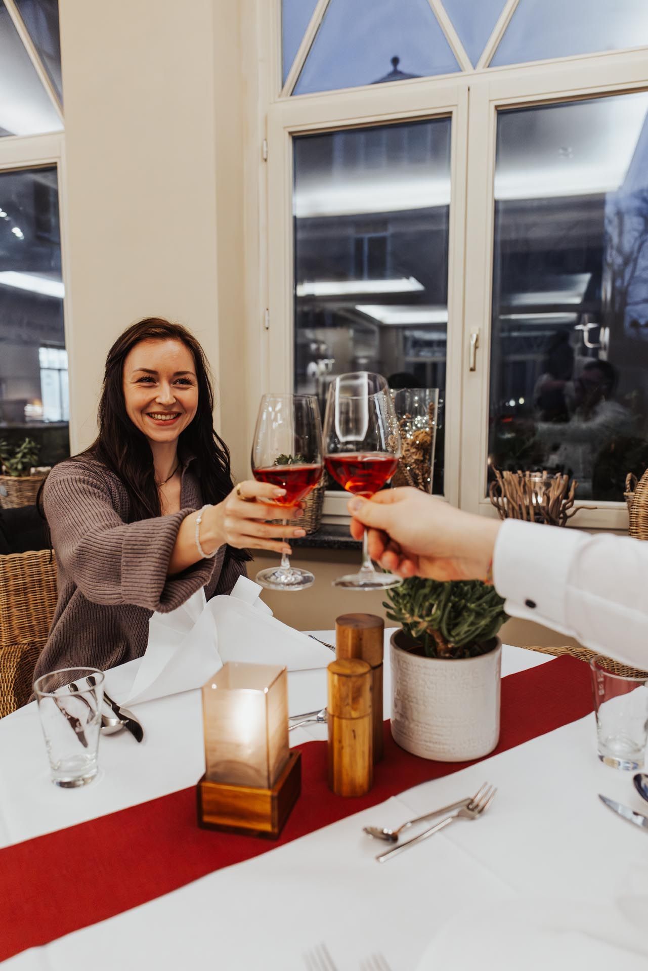 Zwei Personen stoßen an einem Kerzenlicht beleuchteten Restauranttisch am Fenster mit Weingläsern an.