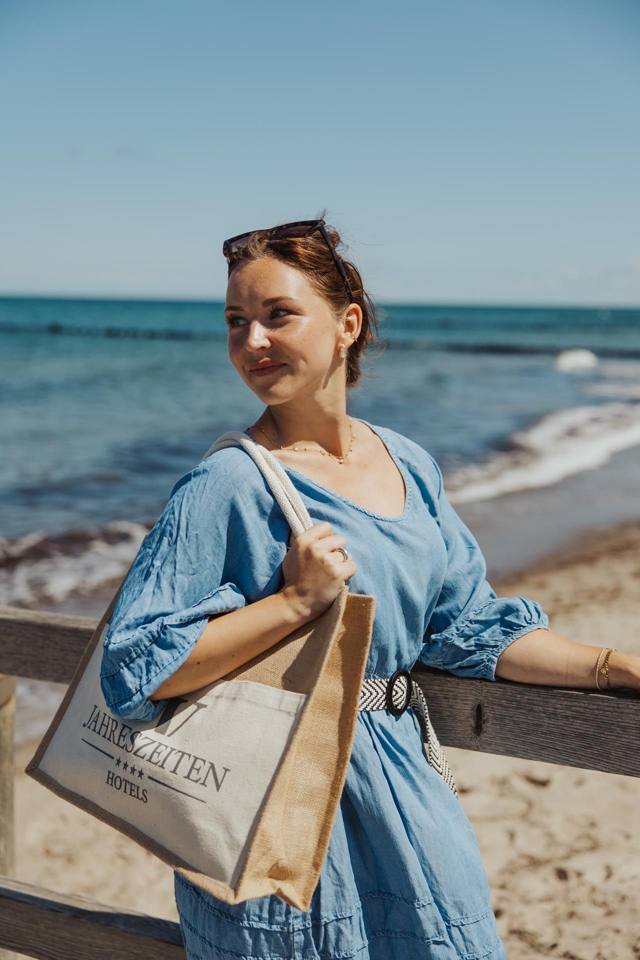 Eine Person mit einer Tragetasche in der Hand steht mit einem blauen Oberteil am Strand neben einem Holzgeländer.