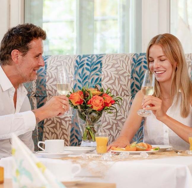 Lächelndes Paar stößt mit Champagner an einem Tisch an. Orange Blumen in einer Vase. Frühstückstisch.