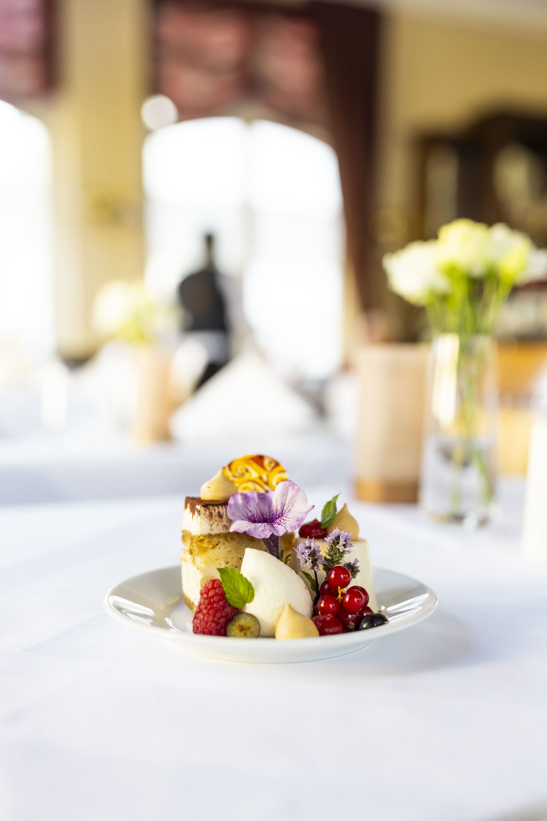 Dessert auf einem weißen Teller: Kuchen, Beeren, Eis und eine Blume; auf einer weißen Tischdecke in einem Esszimmer.