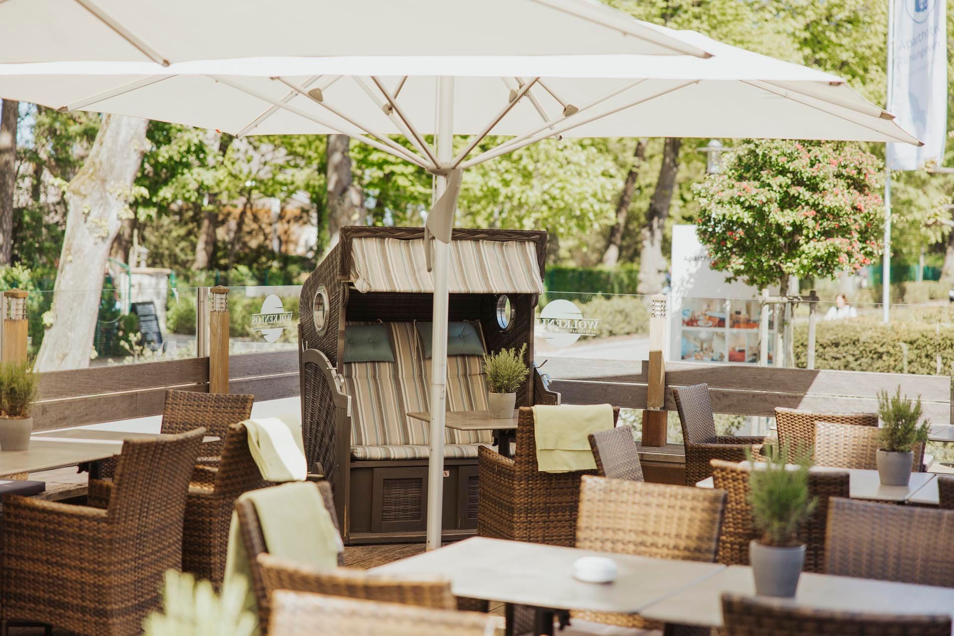 Eine Terrasse mit Tischen, Stühlen und einem Strandkorb unter einem Sonnenschirm.