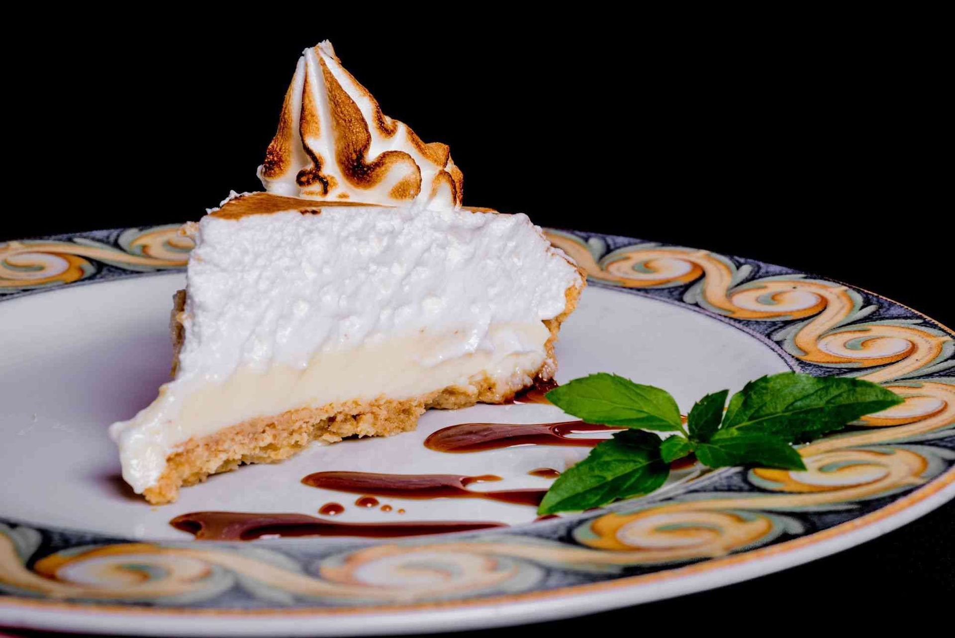 Una rebanada de tarta de merengue de limón en un plato con hojas de menta y salsa de chocolate.
