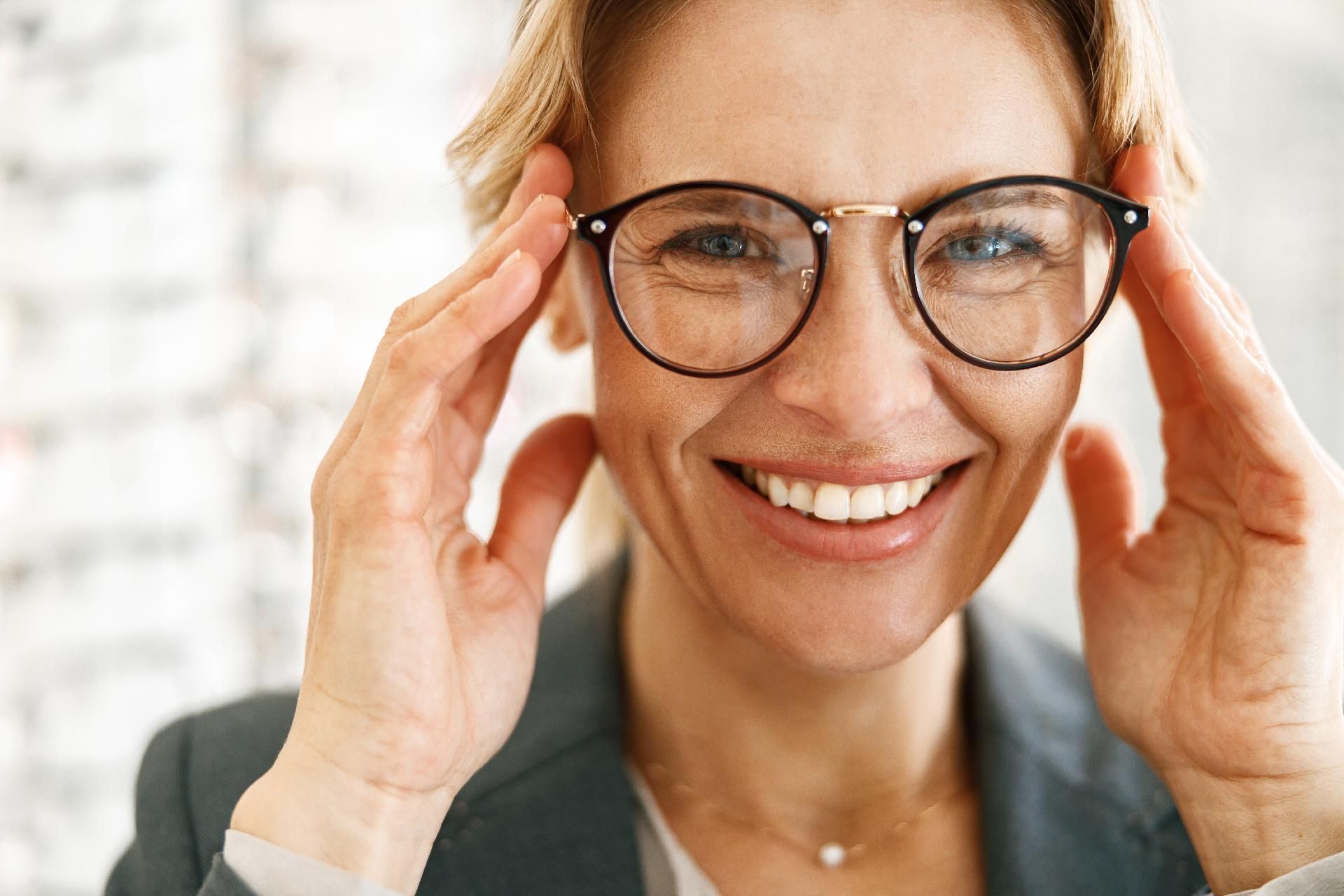 Eine lächelnde Person rückt ihre runde Brille in einer hellen Innenraumumgebung zurecht.
