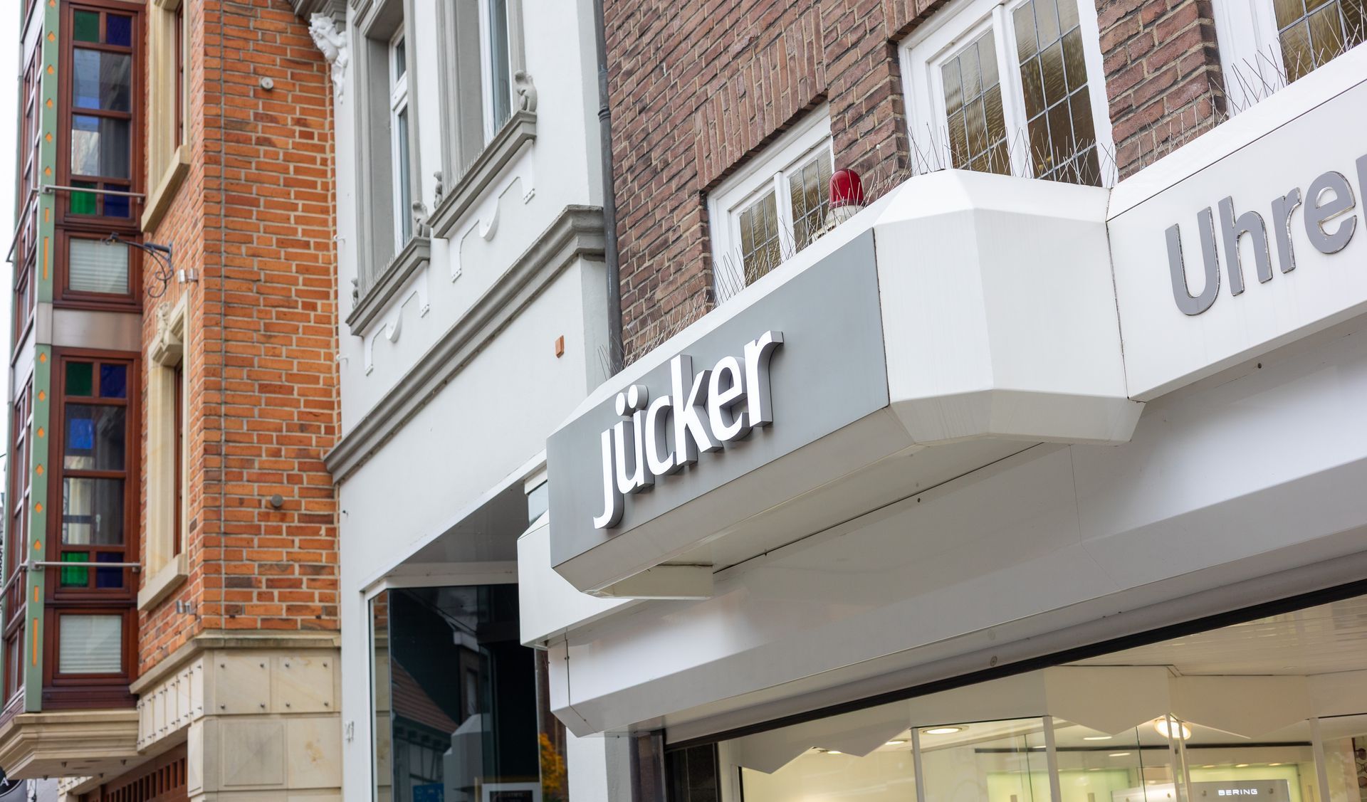 Straßenansicht eines Backsteingeschäfts mit weißem „Jücker“-Schild und Schaufenstern unterhalb von Balkonen