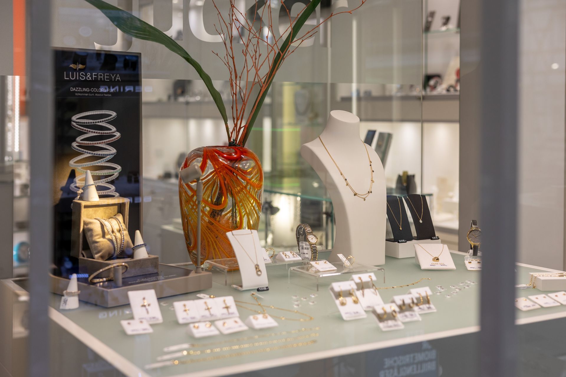 Schmuckauslage in einem Schaufenster mit Halsketten, Ringen und dekorativen Vasen auf einem Glasregal