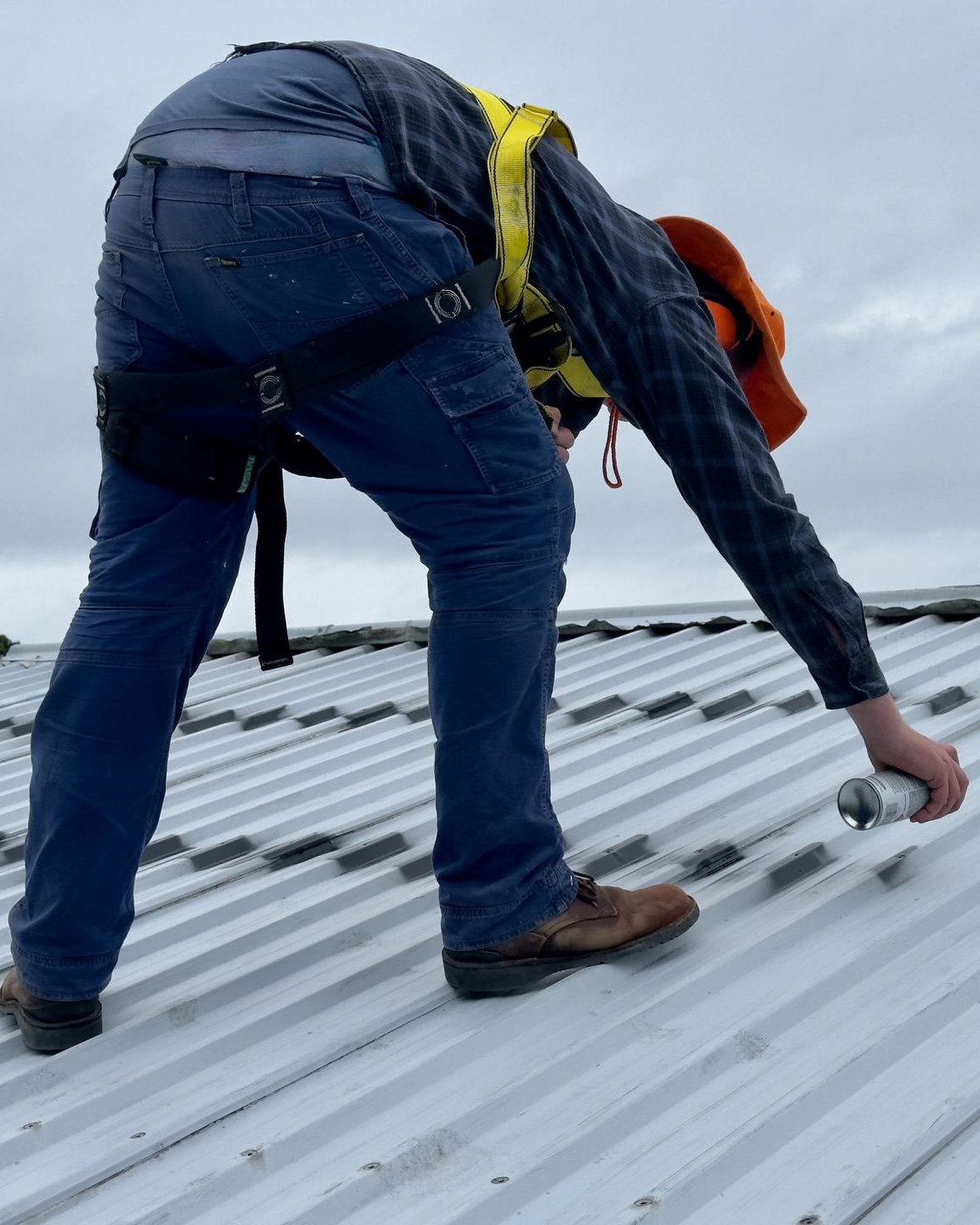 Une personne harnachée pulvérise un produit à l'aide d'une bombe aérosol sur un toit en tôle ondulée. Ciel nuageux.