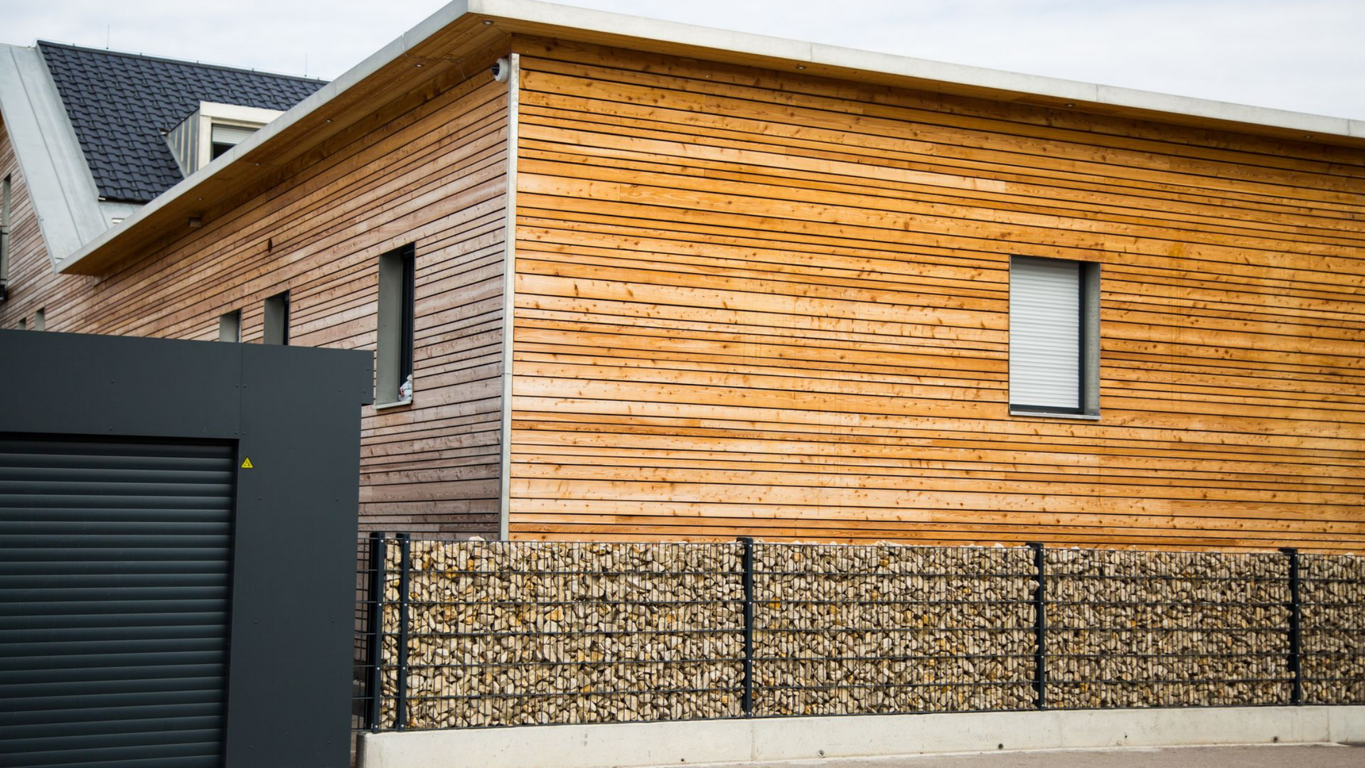 Maison moderne avec bardage en bois, petites fenêtres et clôture en gabions. Porte de garage grise visible.