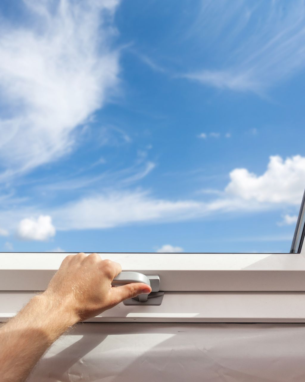 Une main ouvre une fenêtre blanche laissant apparaître un ciel bleu et de fins nuages.