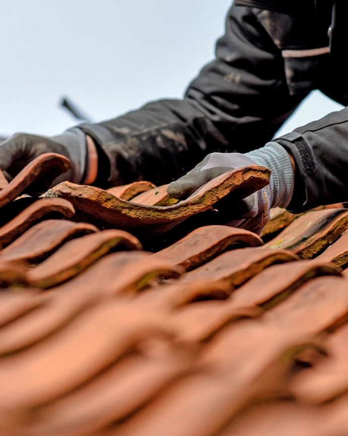 Un couvreur, mains gantées, pose des tuiles de terre cuite rouge sur un ciel bleu.