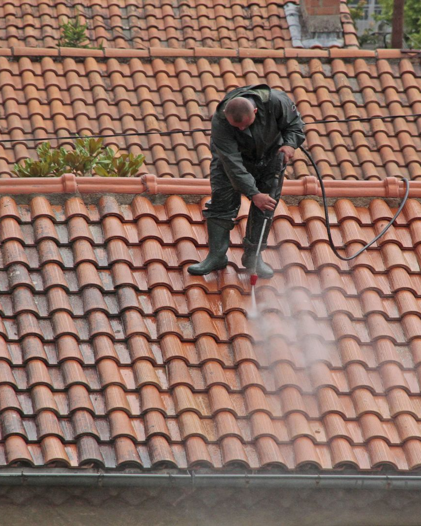 Un homme en costume sombre nettoie au jet d'eau un toit de tuiles rouges.