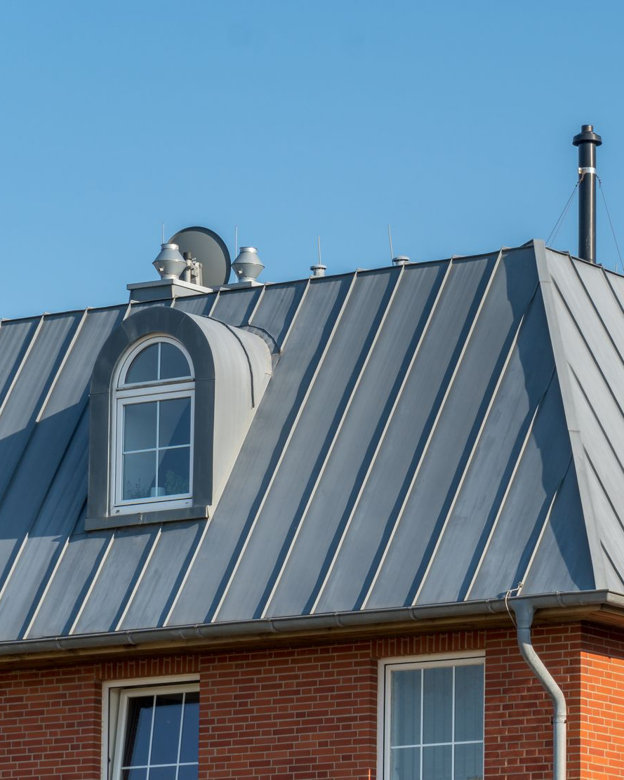 Toiture métallique grise avec lucarne, cheminée et ciel. Bâtiment en briques avec gouttière.