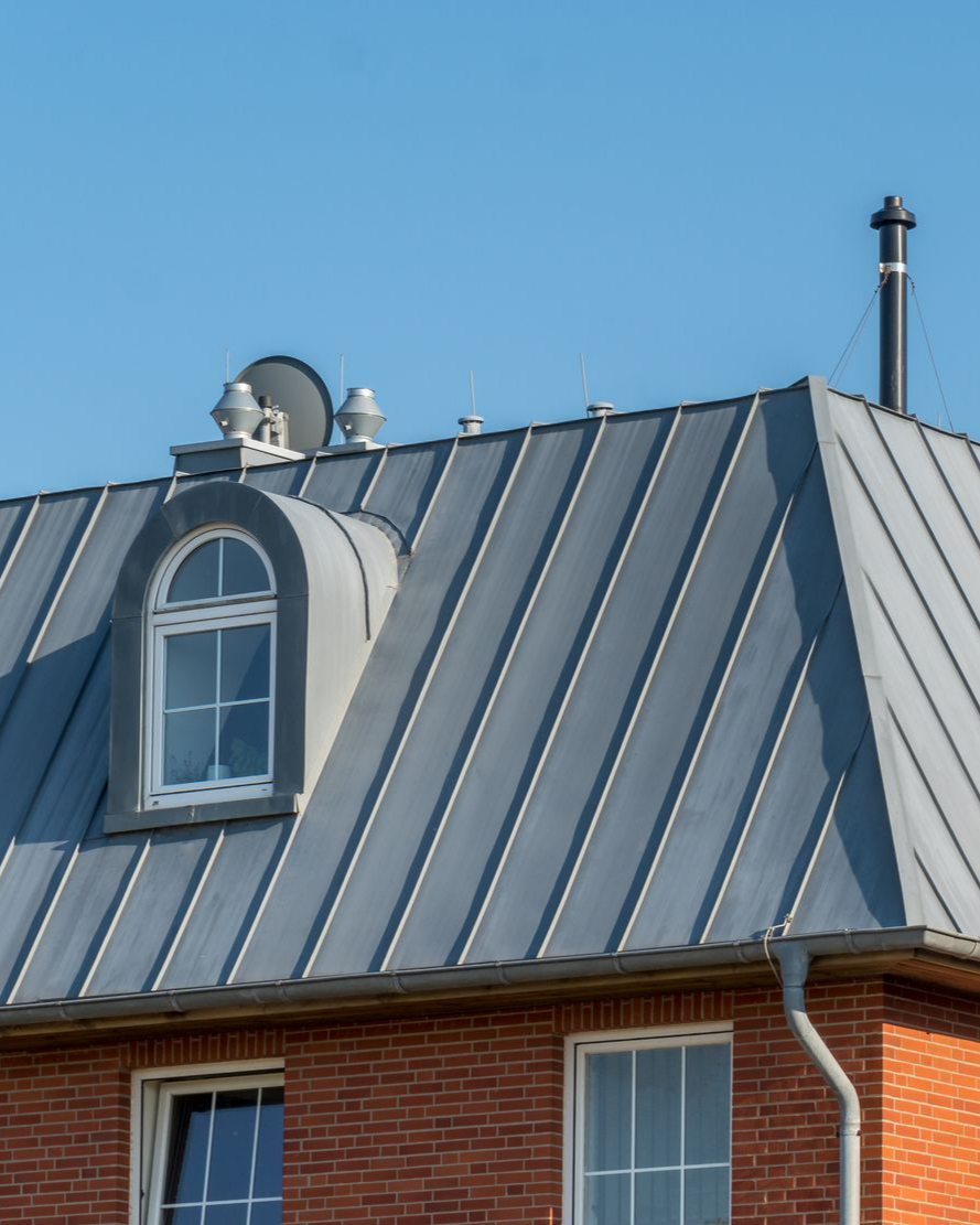 Toiture métallique grise avec lucarne, cheminée et antenne parabolique sur un bâtiment en briques, le tout se détachant sur un ciel bleu.