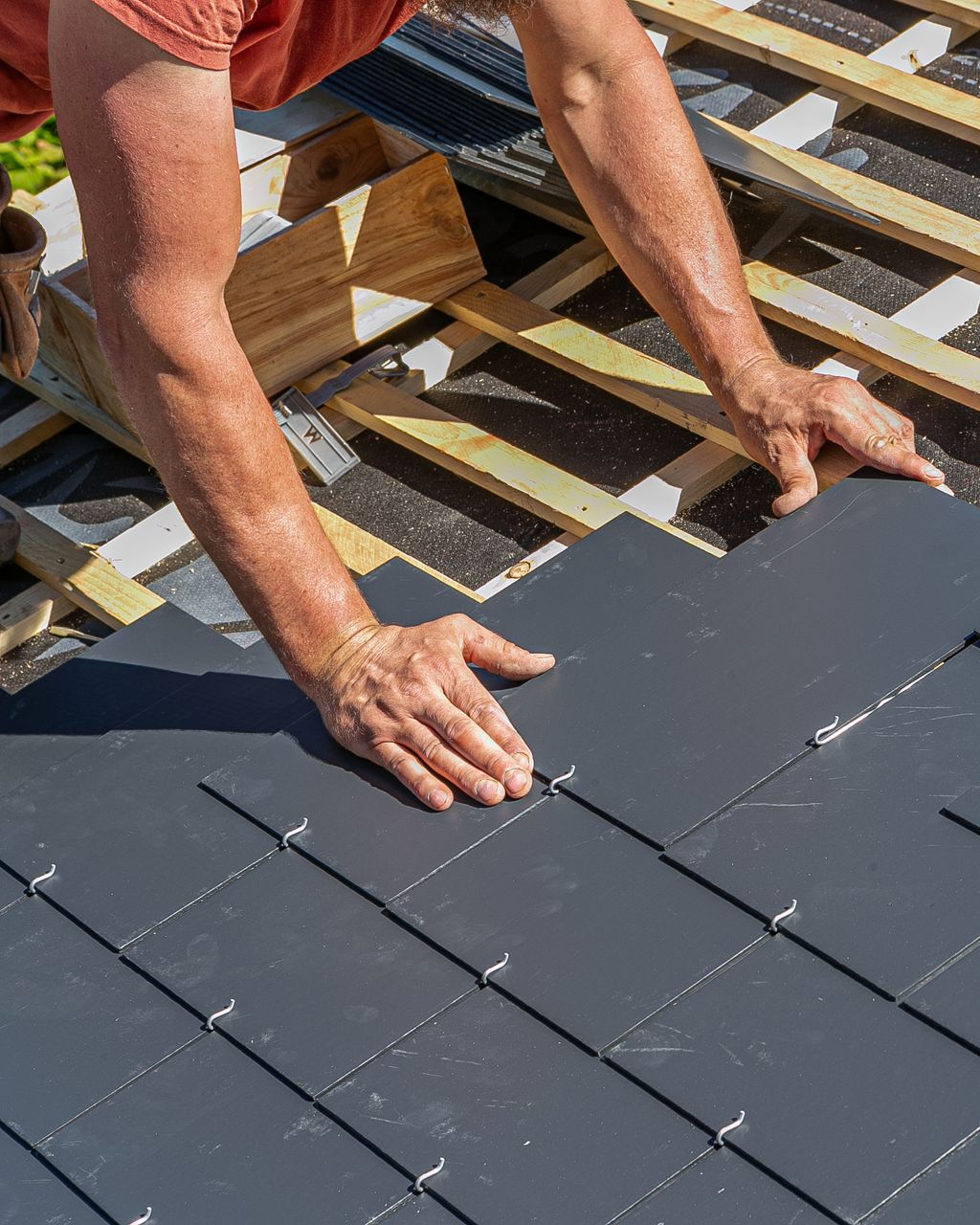 Des mains posent des tuiles de toiture gris foncé sur des chevrons en bois.