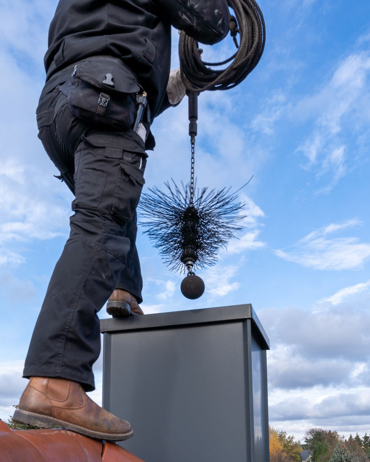 Ramoneur sur un toit, tenant une brosse et un câble, nettoyant une cheminée sur fond de ciel bleu.