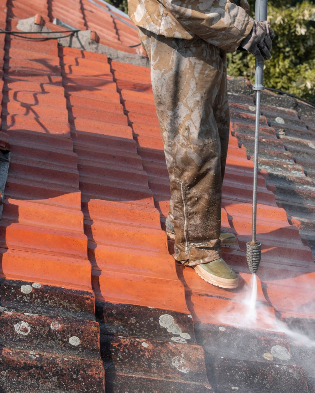 Une personne nettoie à haute pression un toit de tuiles rouges, enlevant la saleté et la mousse.