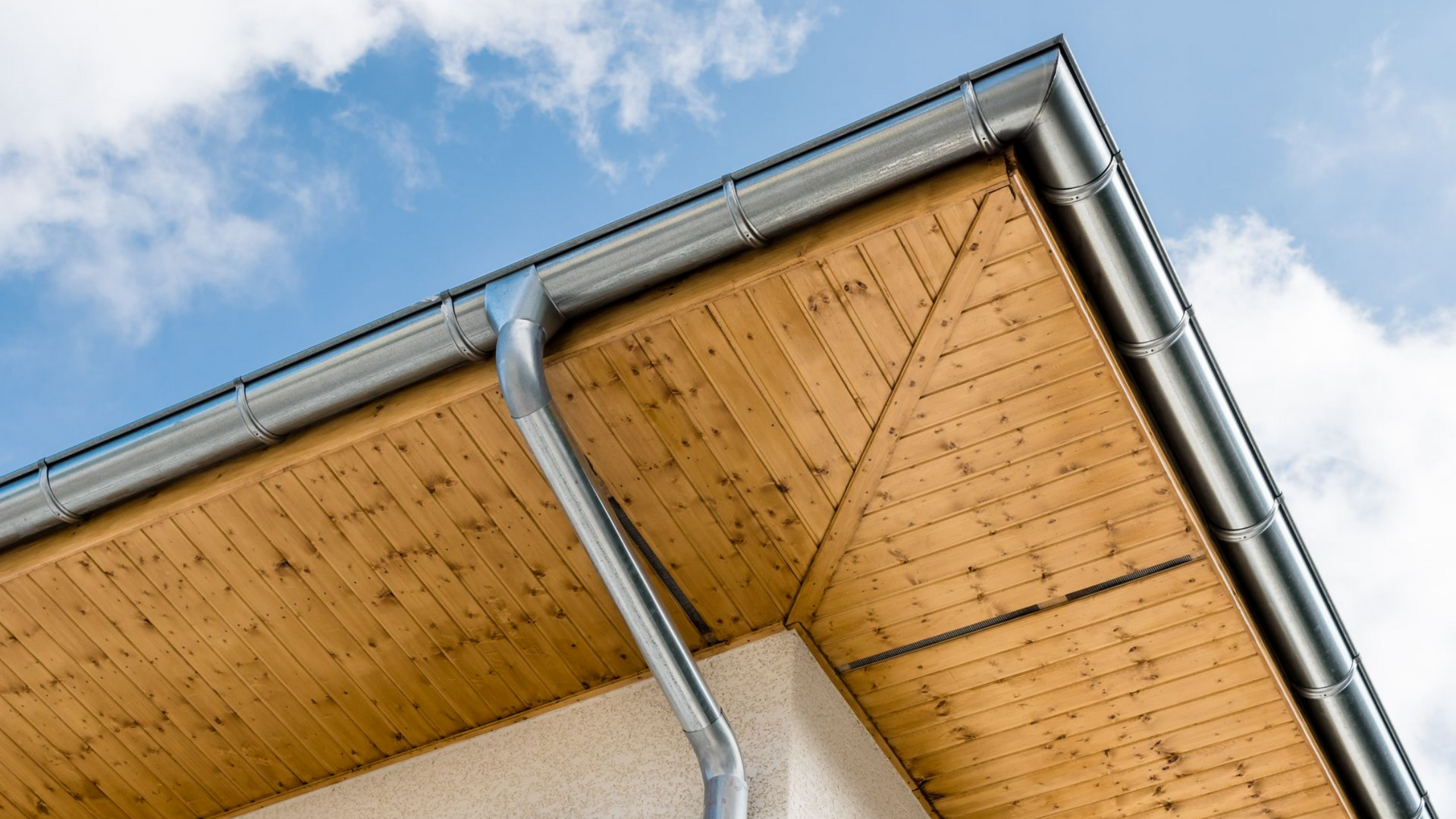 Vue extérieure d'un angle de bâtiment avec un soffite en bois, des gouttières grises et un ciel bleu nuageux.