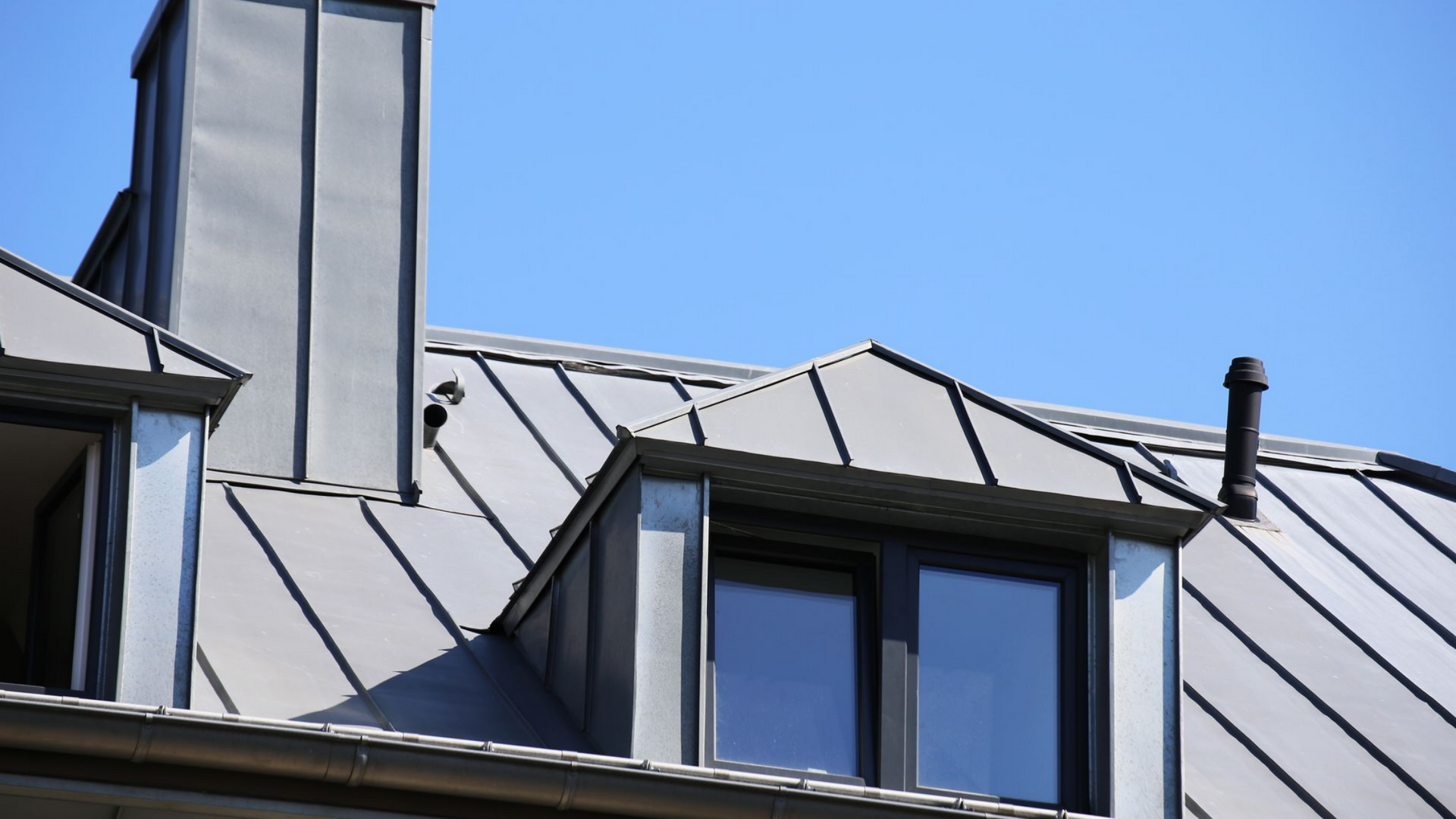 Toiture métallique grise avec lucarne et cheminée sur fond de ciel bleu clair.