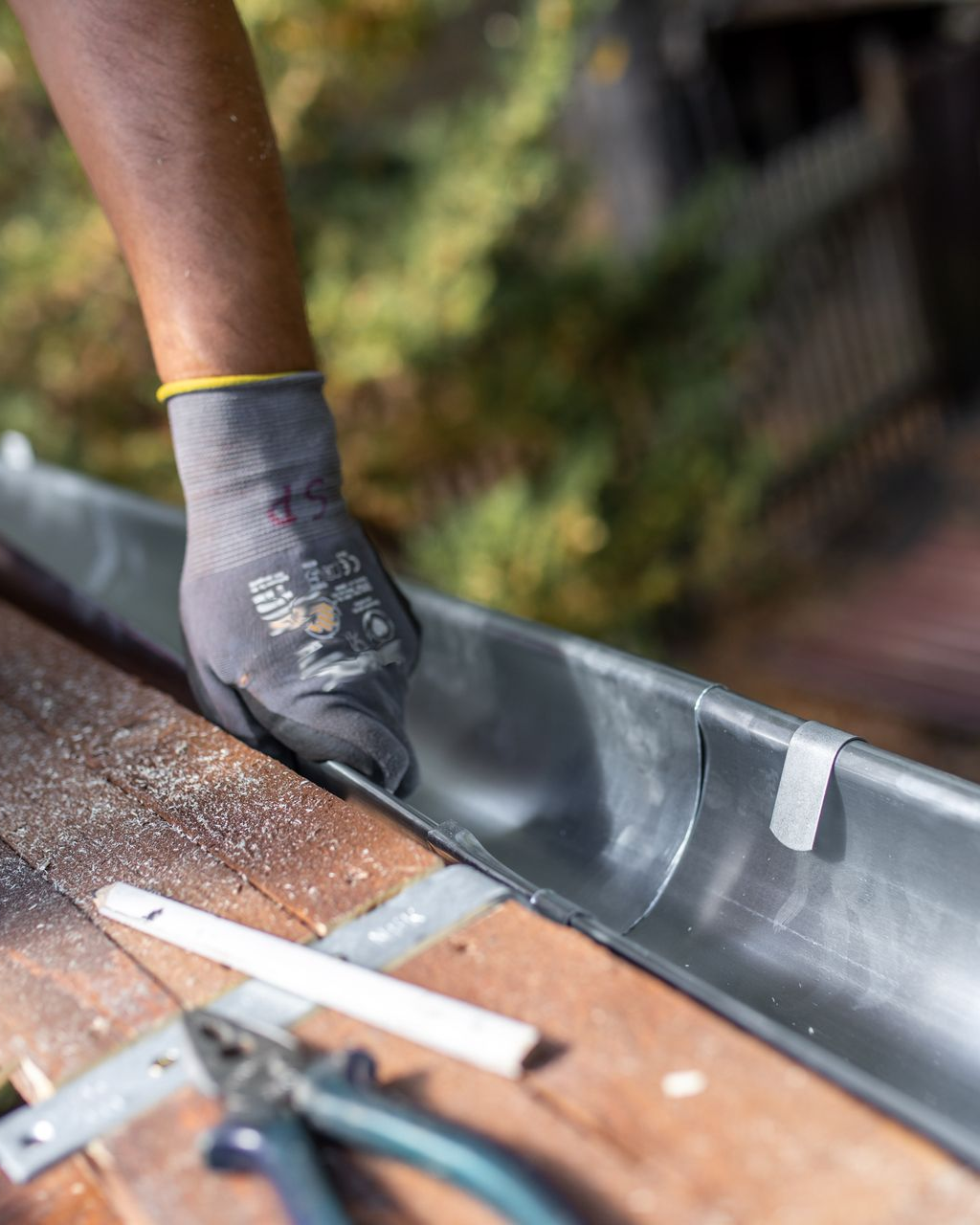 Une personne gantée ajuste une gouttière métallique sur une surface en bois. Des outils et du ruban adhésif sont visibles.