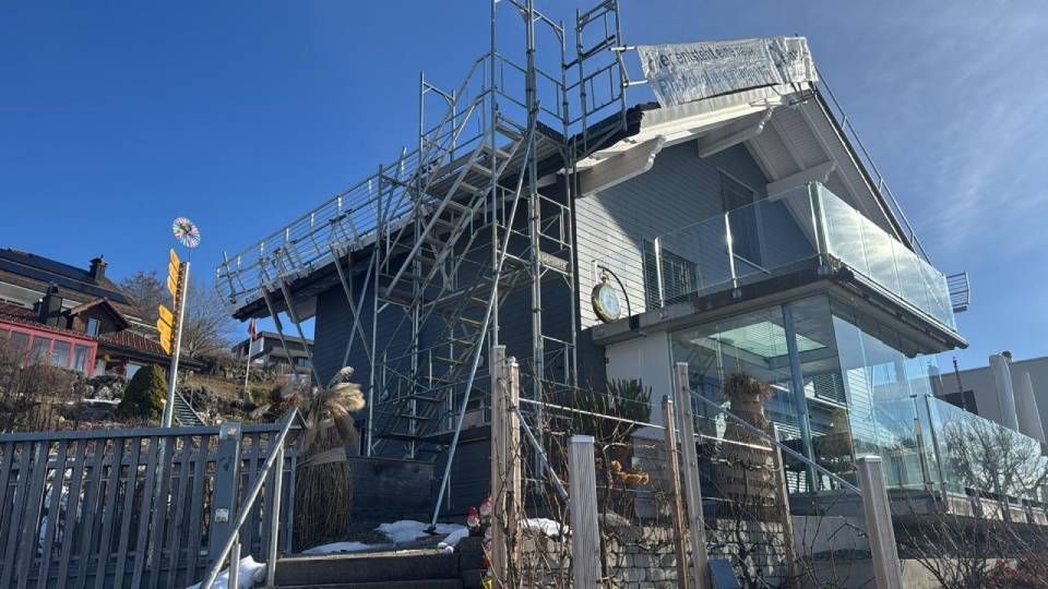 Gerüst auf einem grauen Hausdach mit Glasbalkon; ein sonniger Tag.