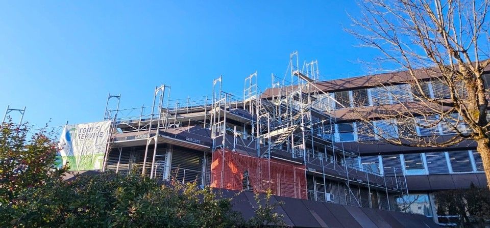 Ein im Bau befindliches Gebäude mit Gerüsten vor blauem Himmel, teilweise verdeckt von kahlen Ästen.