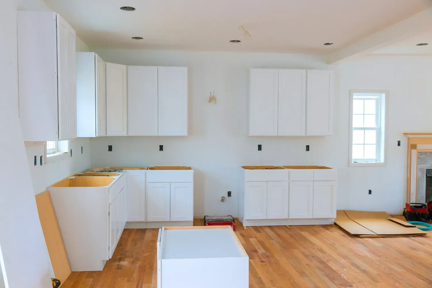 Interior de cocina en construcción con gabinetes blancos, piso de madera clara y paredes sin terminar.