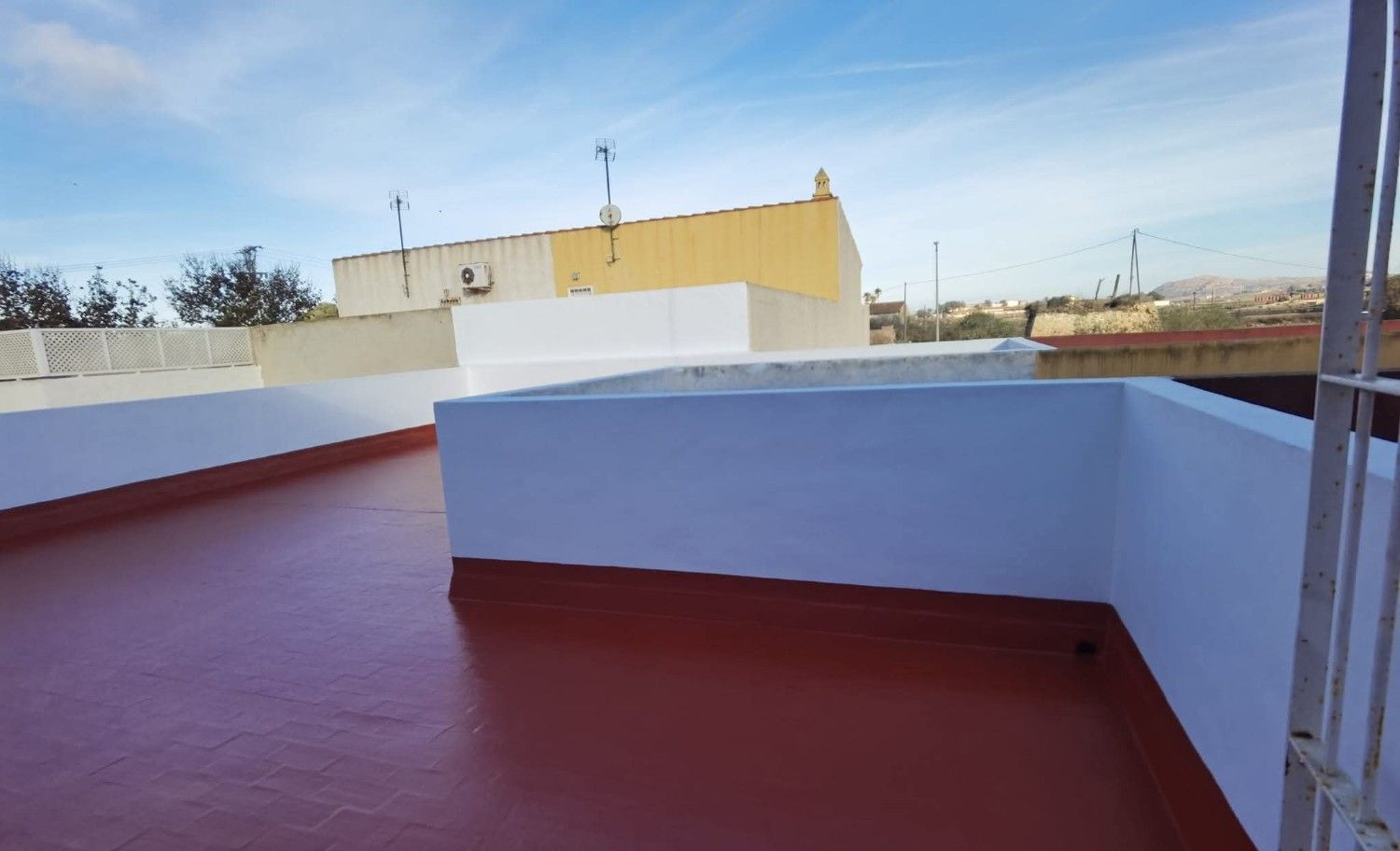 Terraza en la azotea con suelo rojo, paredes blancas y un edificio amarillo al fondo.