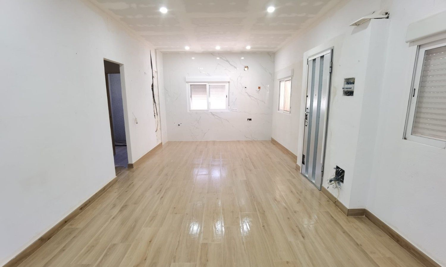 Habitación luminosa y vacía con suelo de madera clara, paredes blancas, luces en el techo y una ventana en la parte trasera.