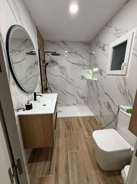 Baño moderno con paredes de mármol, suelo de madera, espejo ovalado, lavabo con mueble, inodoro, ducha y tragaluz.