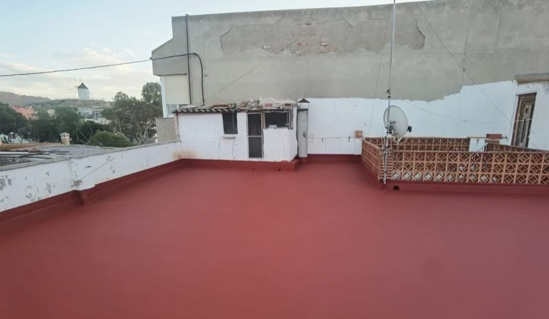 Terraza en la azotea con suelo rojo, paredes blancas y sillas de mimbre junto a una pequeña estructura de servicio.