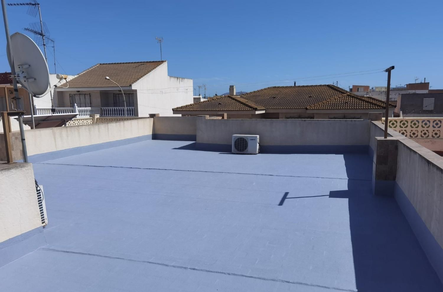 Terraza en la azotea con suelo azul impermeable, muros bajos, antena parabólica y aire acondicionado bajo un cielo despejado.