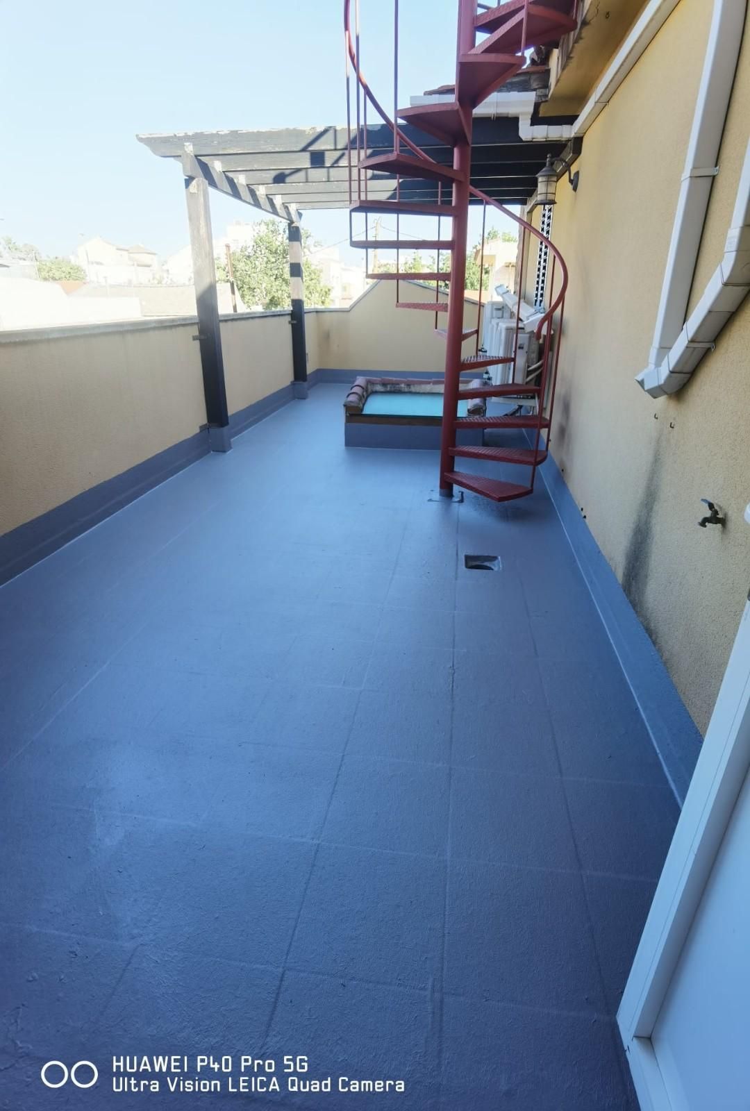 Terraza en la azotea con suelo de baldosas azules, paredes beige y una escalera de caracol roja bajo un toldo metálico.