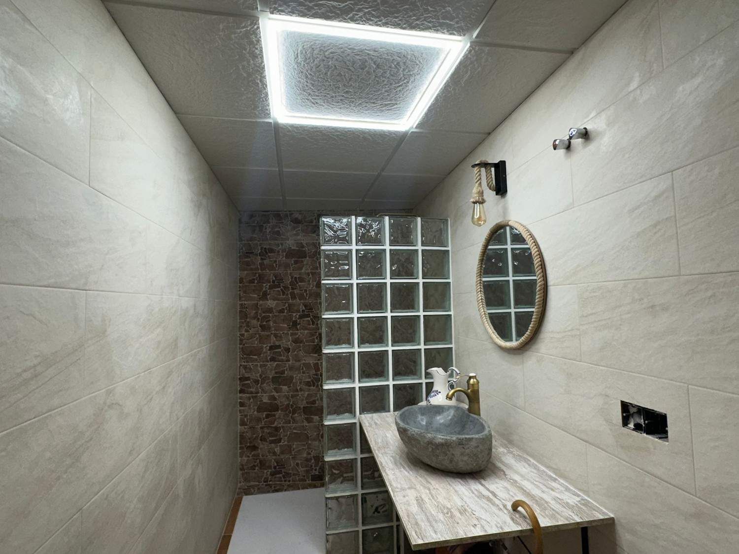 Baño moderno con pared de bloques de vidrio, lavabo de piedra, espejo ovalado y luz de techo brillante.