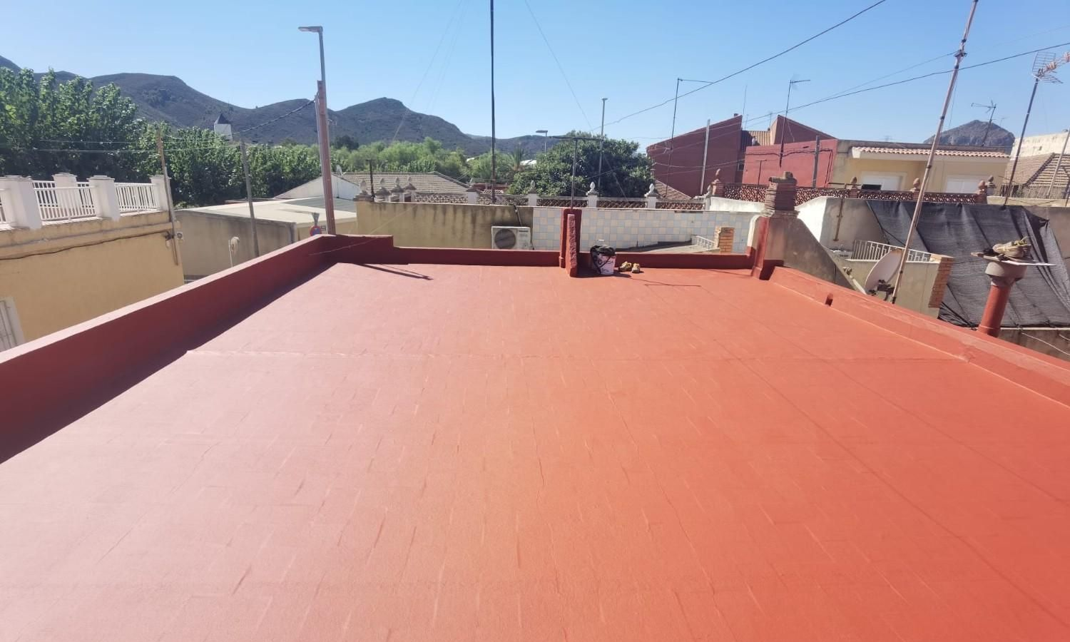 Terraza roja en la azotea con barandillas, antenas y colinas distantes bajo un cielo azul despejado.
