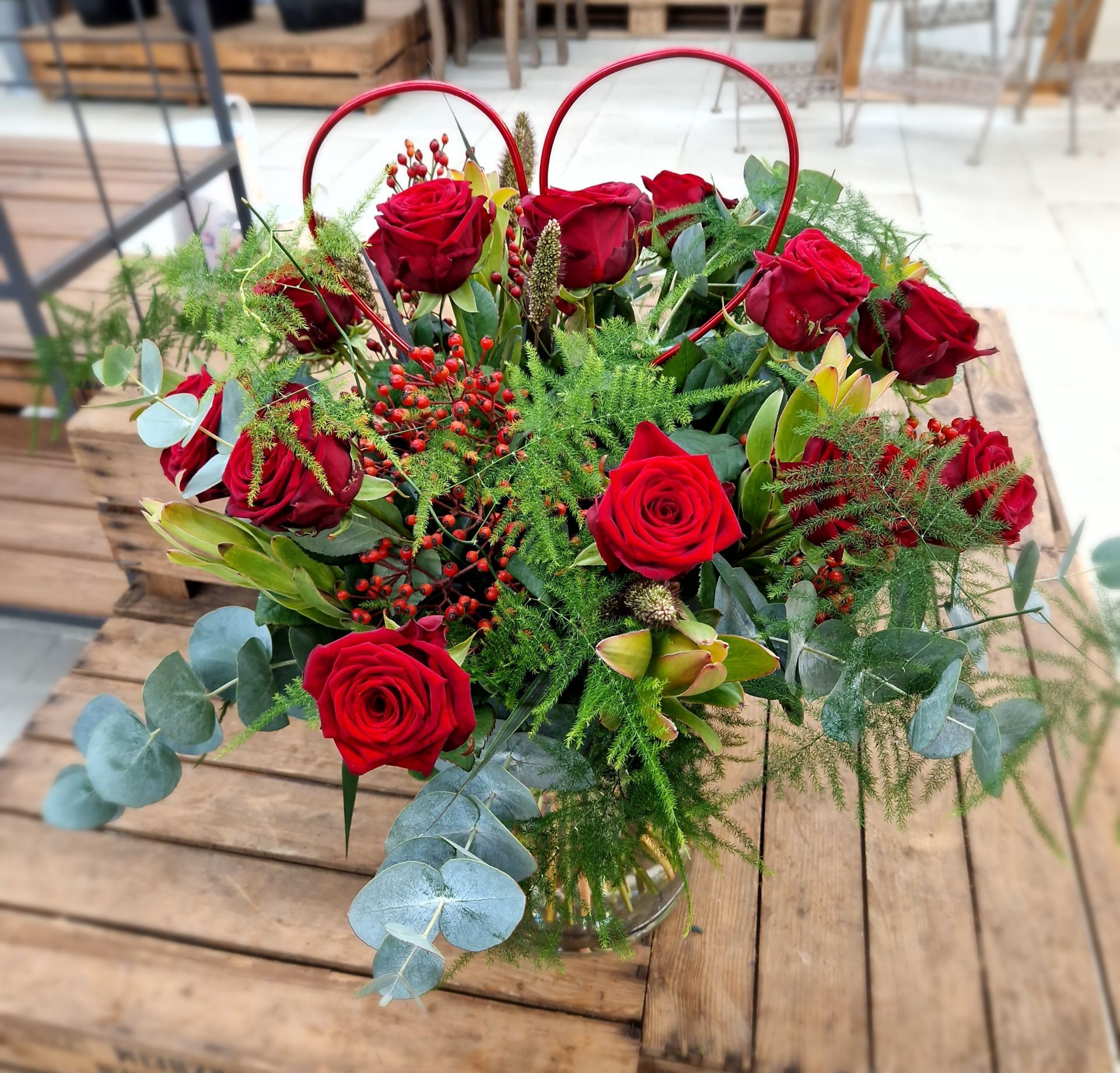 Auf einem Holztisch steht eine Vase mit roten Rosen und Grünpflanzen.